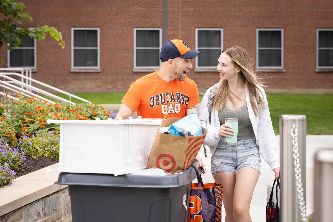Father and daughter smile at each other while moving a bin of belongings into a dorm on move-in day.